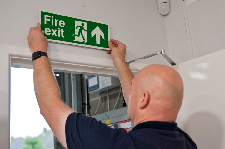 Fire safety signs: engineer with his back to the camera fitting a fire safety sign above a door.